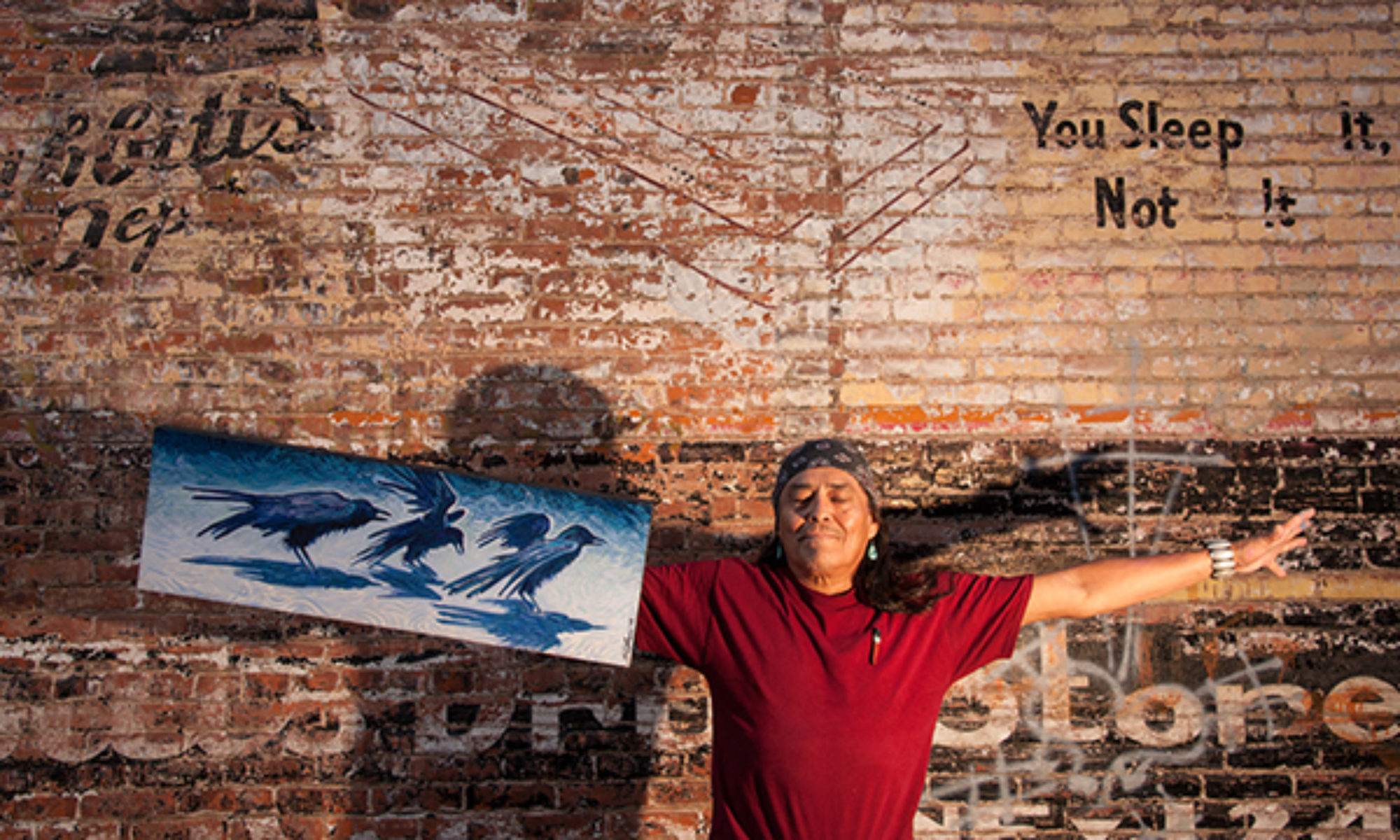 Shonto Begay standing in front of brick wall with arms spread holding one of his paintings. 